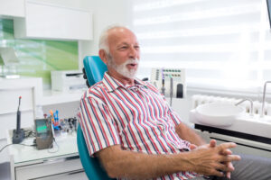 An older man with white hair and a beard, wearing a striped shirt, sits in a dental chair smiling. Dental tools and equipment are on the counter in a bright, modern clinic.