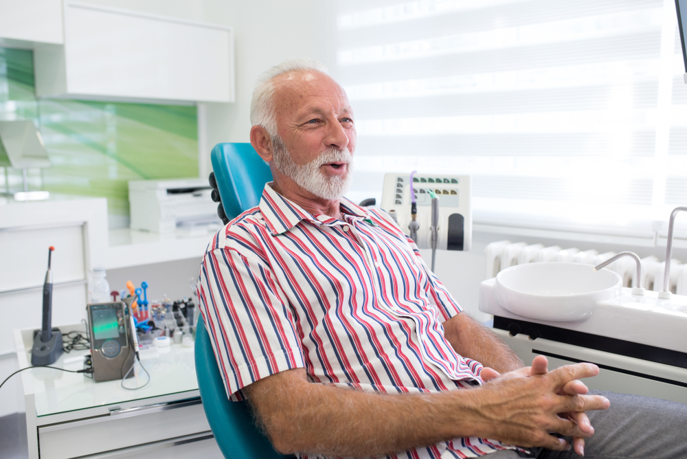 An older man with white hair and a beard, wearing a striped shirt, sits in a dental chair smiling. Dental tools and equipment are on the counter in a bright, modern clinic.