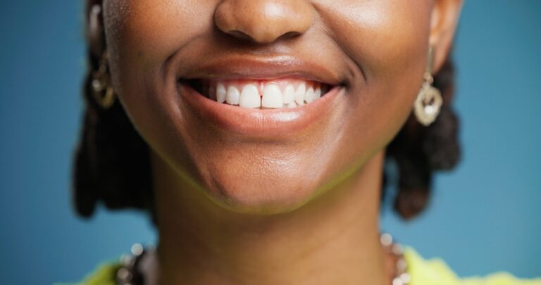 A close-up of a smiling person wearing gold earrings and a necklace against a blue background. Only the lower half of their face is visible, showing white teeth and dimpled cheeks.