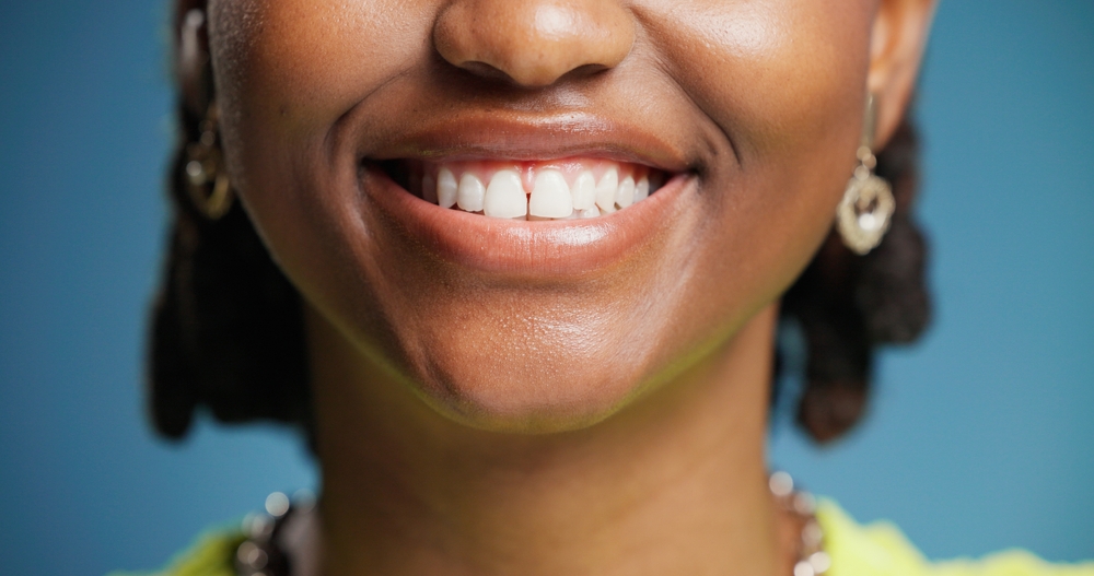 A close-up of a smiling person wearing gold earrings and a necklace against a blue background. Only the lower half of their face is visible, showing white teeth and dimpled cheeks.
